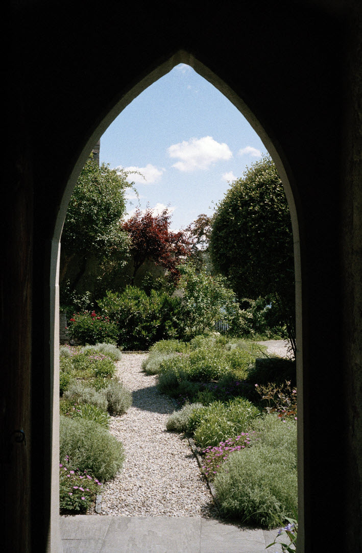 View through a door with a gothic pointed arch into the open. A gravel path leads from the door to a garden. On both sides of the path are cushion plants, some of them with bright pink blooms. There are bushes in the background. The sky above is blue with some white clouds.